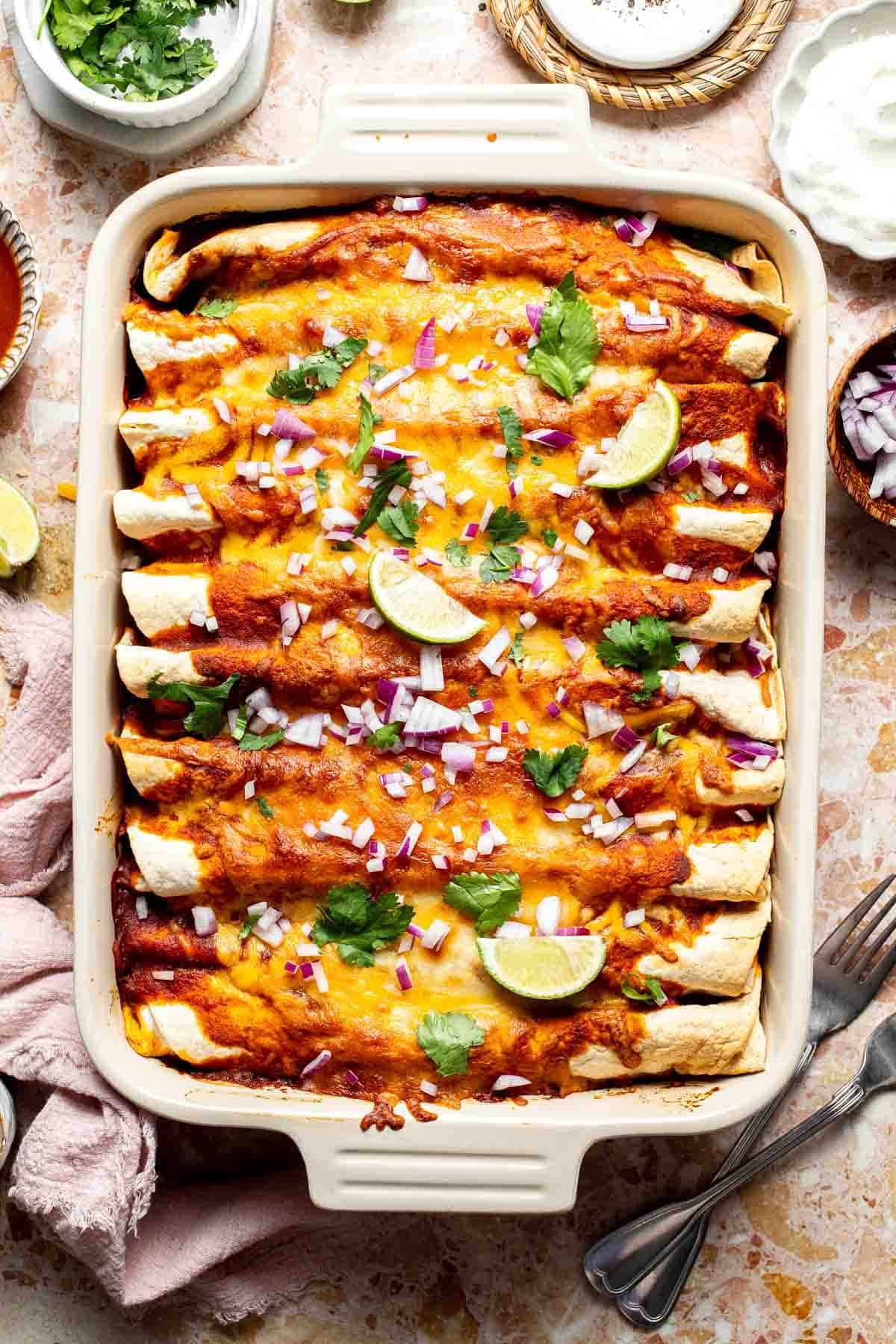Overhead view of a baking dish of sweet potato black bean enchiladas topped with red onion, cilantro, and lime wedges.
