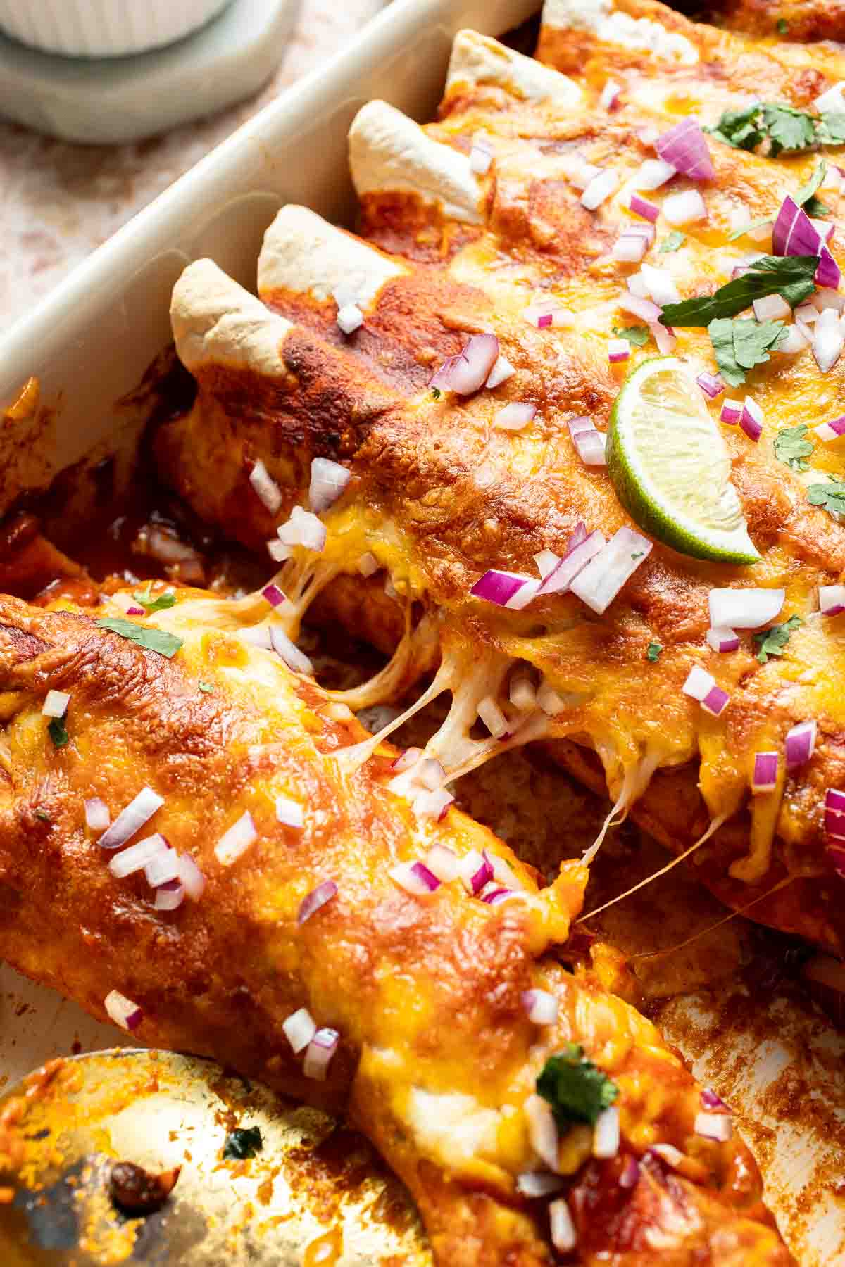 Close-up of cheesy baked sweet potato black bean enchiladas topped with melted cheese, red onion, cilantro, and lime wedges.
