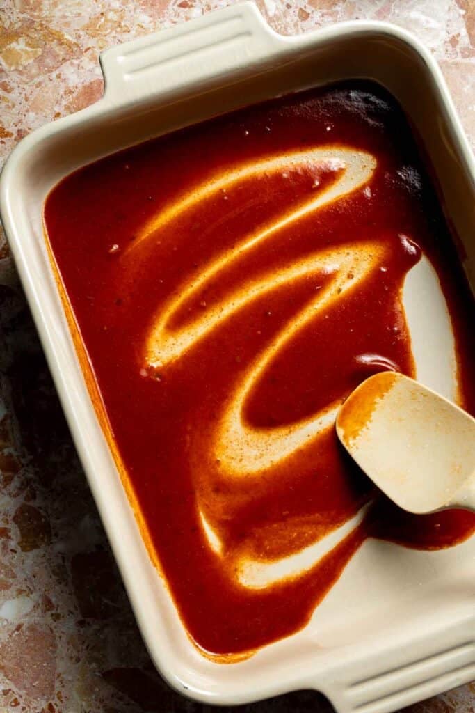Red enchilada sauce spread across the bottom of a baking dish.