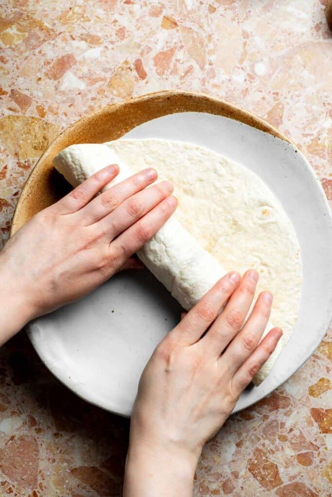 Hands rolling a flour tortilla tightly around the sweet potato filling.