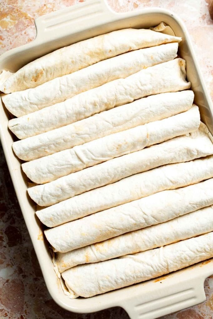 Rolled enchiladas lined up seam-side down in a baking dish.