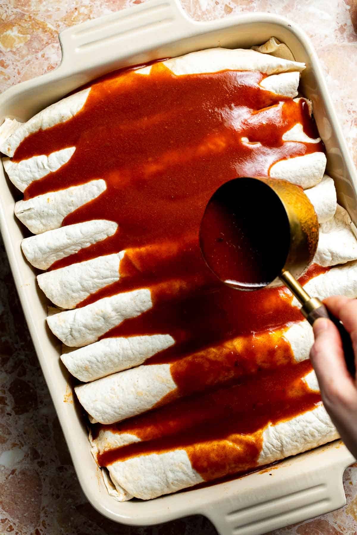 Pouring red enchilada sauce over the rolled enchiladas in the baking dish.