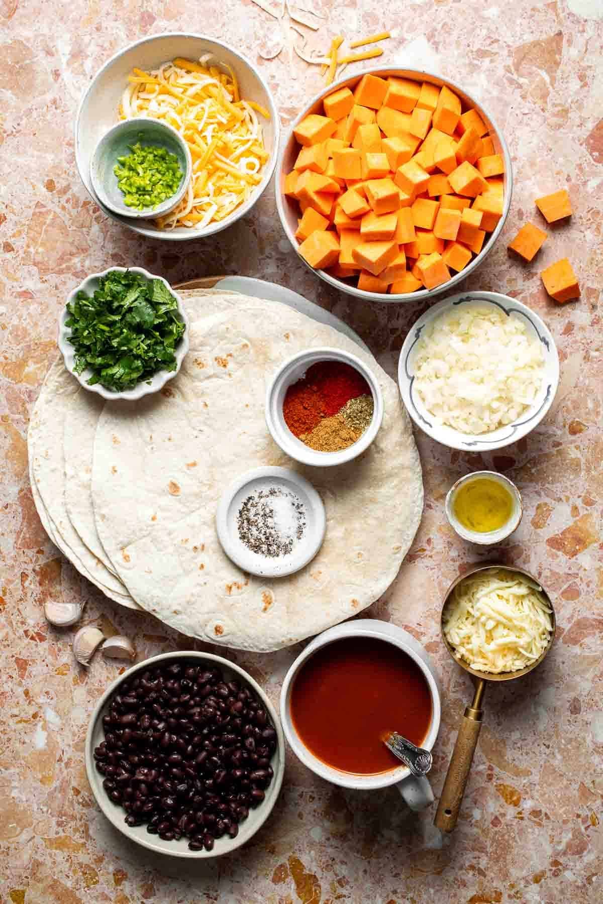 Ingredients for sweet potato black bean enchiladas, including diced sweet potatoes, black beans, flour tortillas, shredded cheese, jalapeño, cilantro, spices, and enchilada sauce, arranged on a pink marble surface.