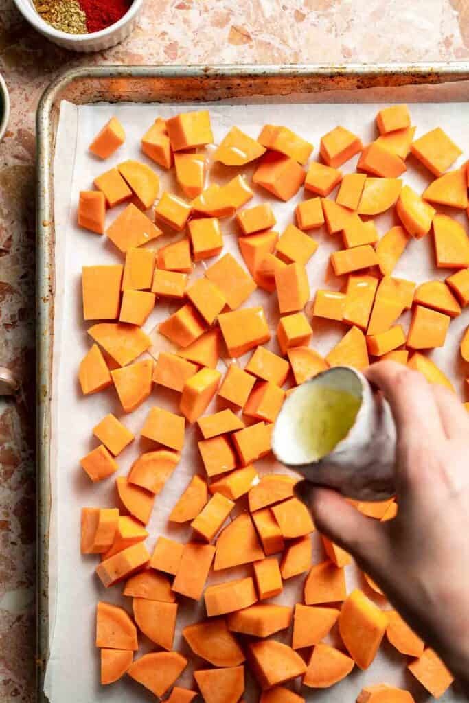 Pouring olive oil over diced sweet potatoes on a parchment-lined baking sheet.