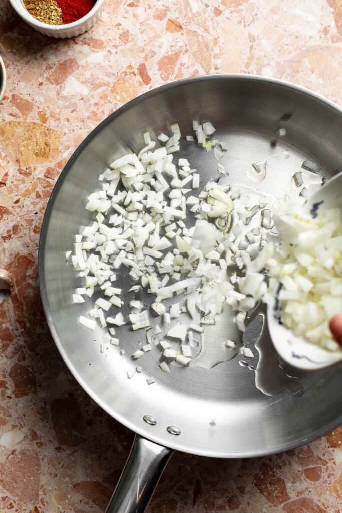 Diced onion being added to a stainless steel skillet.