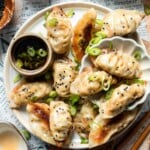 Vegetable gyoza on a plate with crispy golden bottoms, black sesame seeds, and sliced green onions, served with a bowl of dipping sauce and chopsticks.