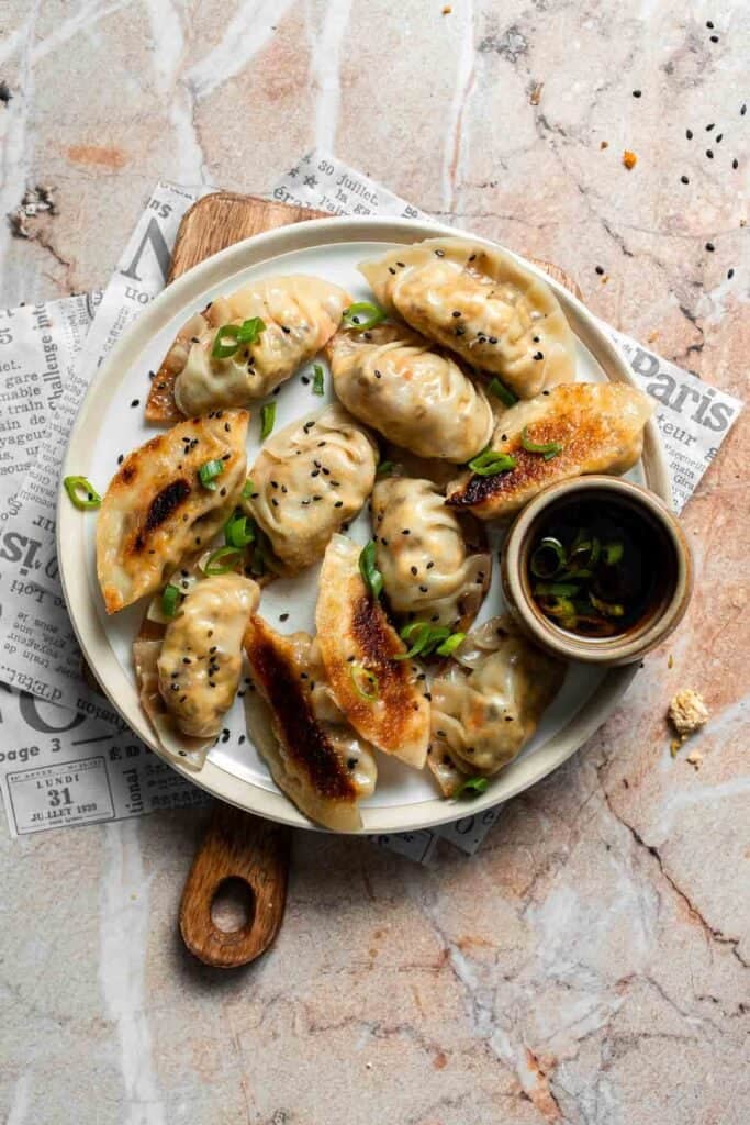 Plate of crispy pan-fried vegetable gyoza topped with green onions and black sesame seeds, with a small bowl of dipping sauce.