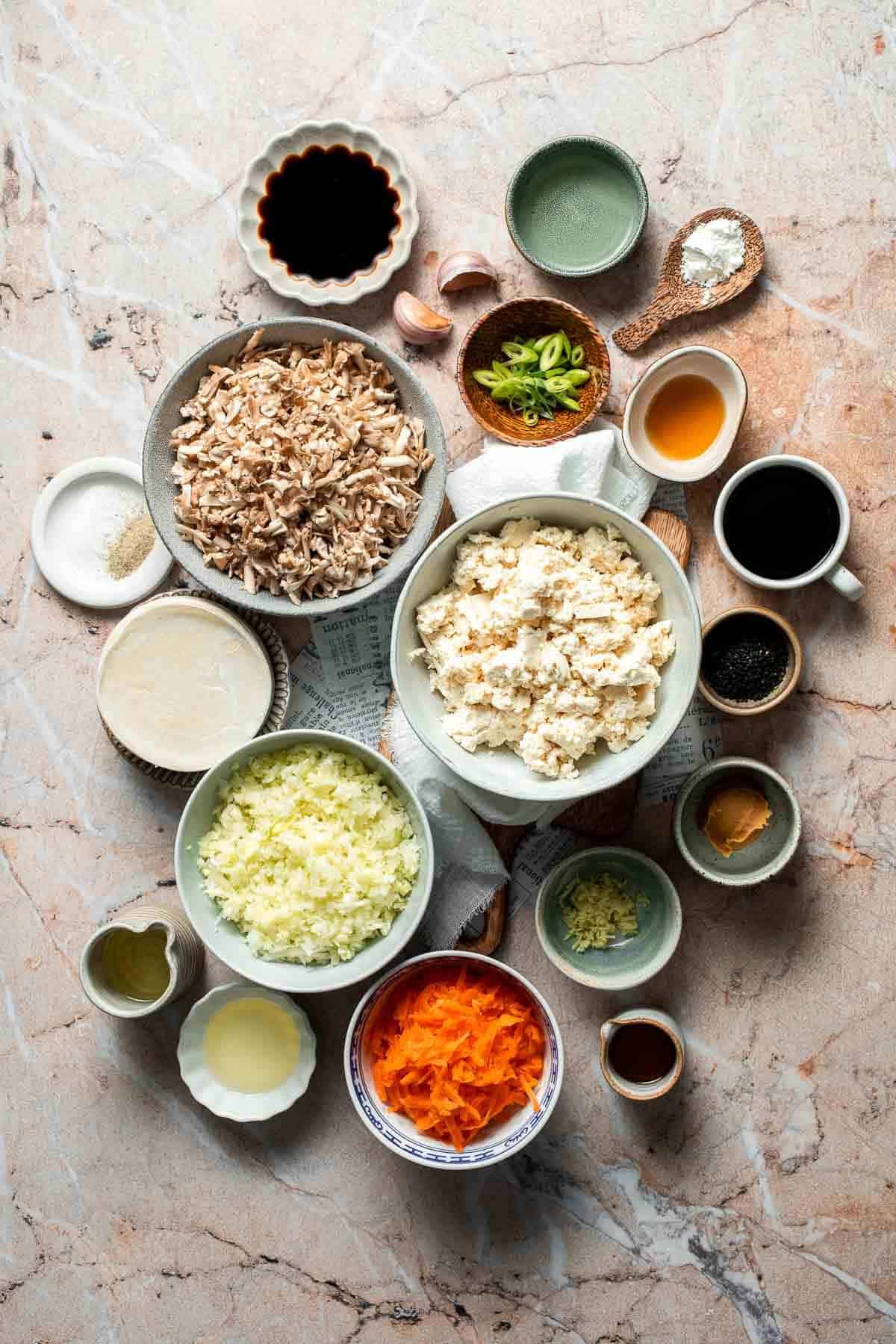 Ingredients for vegetable gyoza, including shredded mushrooms, crumbled tofu, shredded cabbage, carrot, miso, soy sauce, sesame oil, garlic, ginger, and green onions, arranged on a marble surface.