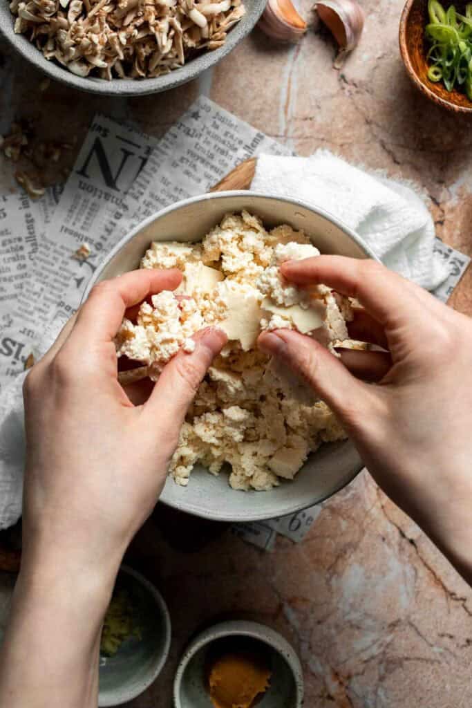 Hands crumbling a block of pressed tofu into a bowl.