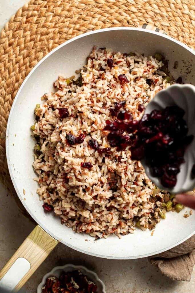 Adding dried cranberries to the wild rice and vegetable mixture in the skillet.