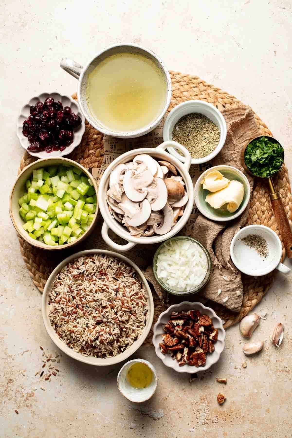 Ingredients for wild rice stuffing, including wild rice blend, vegetable broth, diced onion and celery, sliced mushrooms, butter, dried cranberries, pecans, parsley, and spices, arranged on a light surface with a woven placemat.